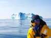 Persona con giacca gialla e occhiali da sole su una barca vicino a un grande iceberg in mare calmo.