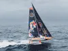 IMOCA foiling con vele scure e scafo colorato che sfreccia sollevato sui foil su un mare grigio, con la scritta 11th Hour Racing