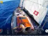 Veliero visto dall'alto in mare aperto, con vela bianca recante una croce svizzera e un velista sul ponte vicino al tettuccio arancione