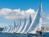Fila di barche a vela J/22 in regata davanti al lungomare di Napoli con cielo sereno.