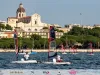 Barche OpenSkiff con vele rosse e nere in regata davanti al lungomare di Cagliari, con una chiesa a cupola sullo sfondo