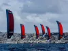 Sei kite rossi e blu in fila sul mare, con atleti su foil in acqua e la città sullo sfondo sotto un cielo nuvoloso