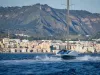Un AC75 bianco e blu naviga in foiling davanti a una costa urbanizzata, con una collina verde sullo sfondo