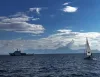 Barca a vela con randa bianca in mare aperto e, sullo sfondo, una nave grigio-bianca sotto un cielo azzurro con nuvole e montagne all'orizzonte