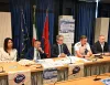 Tavolo con cinque persone sedute davanti a microfoni durante una conferenza stampa, bandiere e poster sullo sfondo