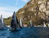 Barche a vela con equipaggi in regata su un lago, sotto una ripida montagna rocciosa e boscosa, con cielo azzurro e qualche nuvola