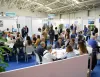 Spazio espositivo affollato con tavoli di lavoro e stand fieristici, persone in discussione e confronti professionali.