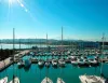 Porto turistico con barche a vela e a motore ormeggiate in acque calme, sole in alto, colline e costa sullo sfondo
