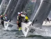 Barche a vela con vele grigie e numeri 122 e 202 navigano vicine in regata su un lago, con gli equipaggi a bordo inclinati sopravvento.