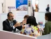 Incontro a un tavolo in fiera: un uomo in giacca gesticola parlando con tre donne, accanto a un cartello con la bandiera sudafricana.