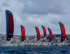 Sei kite rossi e blu in fila sul mare, con atleti su foil in acqua e la città sullo sfondo sotto un cielo nuvoloso