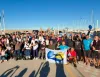 Partecipanti e organizzatori sul molo di un porto che tengono una bandiera con logo, imbarcazioni alle spalle