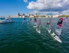 Piccole derive con vele rosse e nere navigano in fila su un mare blu davanti a una città costiera, con una barca a motore bianca sulla sinistra