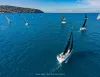 Veduta aerea di una flotta di barche a vela con vele nere che navigano su un mare blu, con la costa collinare sullo sfondo.