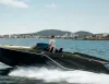 Imbarcazione sportiva di lusso scura in navigazione con un uomo al timone e la costa sullo sfondo