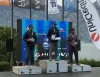 Tre uomini sul podio della premiazione davanti al lago, con bandiere e pannello blu sullo sfondo; quello al centro regge un trofeo