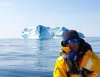Persona con giacca gialla e occhiali da sole su una barca vicino a un grande iceberg in mare calmo.