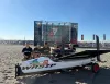 Barca WASZP sulla spiaggia con un gruppo di persone e un palco del Race Village sullo sfondo