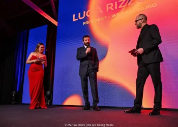 Tre persone su un palco durante un evento, un uomo parla al microfono davanti a uno schermo luminoso.