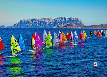 Flotta di windsurfer con vele colorate sul mare davanti a un costone roccioso