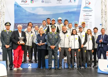 Gruppo di velisti in giacca chiara e autorità in divisa posano sul podio con trofei davanti al pannello del Campionato Italiano Classi Olimpiche