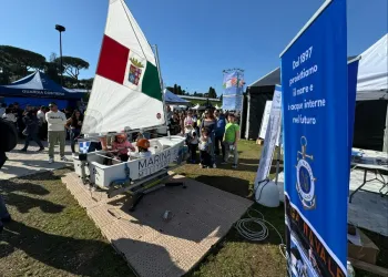 Simulatore di Optimist con vela tricolore su pedana, bambini a bordo e visitatori intorno, accanto a un banner blu della Lega Navale