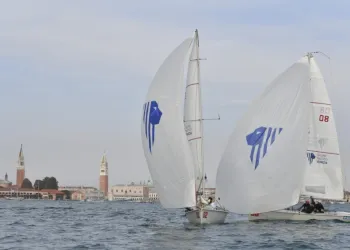 Due barche a vela con spinnaker bianchi e logo blu navigano in laguna, con il campanile di San Marco e San Giorgio Maggiore sullo sfondo.