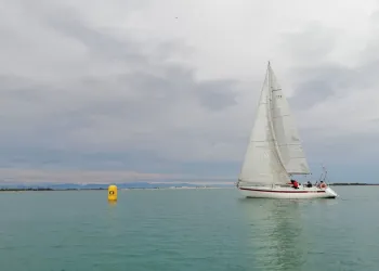 Barca a vela bianca con vele quasi flosce su mare piatto, accanto a una boa gialla, sotto un cielo coperto