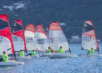 Flotta di derive O'pen Skiff con vele rosse e grigie e numeri velici, timonieri in maglia giallo fluo, davanti a una costa verde