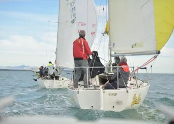 Due J24 in regata con equipaggi a bordo, vele bianche e spinnaker giallo, mare mosso e costa sullo sfondo