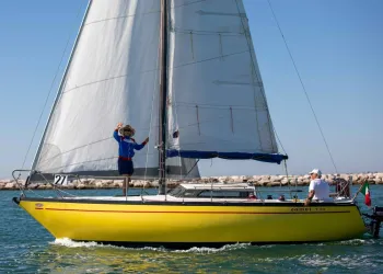Imbarcazione a vela gialla in regata con equipaggio e vele spiegate davanti a un molo