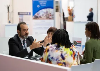 Incontro a un tavolo in fiera: un uomo in giacca gesticola parlando con tre donne, accanto a un cartello con la bandiera sudafricana.