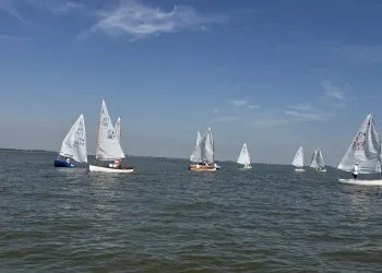 Dinghy 12' con vele bianche in acqua aperta sotto un cielo azzurro, con barche sparse sull'orizzonte e mare leggermente increspato