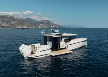 Barca a motore bianca con due fuoribordo e bandiera finlandese in navigazione su mare calmo, con costa montuosa sullo sfondo