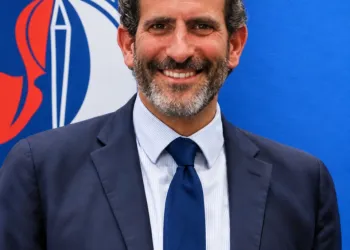 Uomo sorridente con barba, giacca e cravatta blu con ancora dorata, davanti a un fondale blu con logo parziale di Assonautica