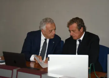 Due uomini in giacca e cravatta discutono documenti seduti a un tavolo con laptop
