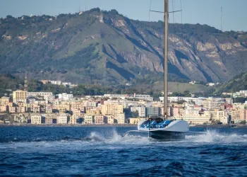 Un AC75 bianco e blu naviga in foiling davanti a una costa urbanizzata, con una collina verde sullo sfondo