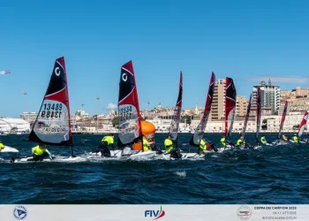 Fila di Open Skiff in regata davanti al lungomare di Cagliari con vele rosse e equipaggi in giubbotti fluorescenti