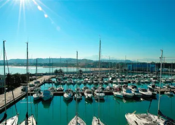Porto turistico con barche a vela e a motore ormeggiate in acque calme, sole in alto, colline e costa sullo sfondo
