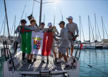 Equipaggio giovane sul ponte di una barca da regata che tiene una bandiera nel porto di Trieste.