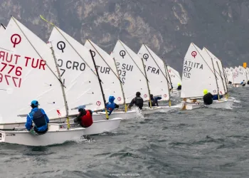 Una lunga fila di Optimist in linea di partenza sull'acqua mossa, con i velisti in assetto e le montagne sullo sfondo.