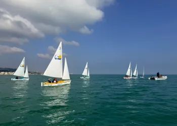Piccole barche a vela con vele bianche navigano su un mare verde e calmo, con un gommone di assistenza a destra e la costa sullo sfondo.