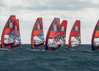Windsurf con vele arancioni e bandiera italiana in fila su mare mosso sotto un cielo nuvoloso