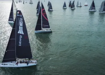 Veduta aerea di una flotta di barche a vela con vele scure in mare calmo; in primo piano uno yacht con la scritta Gruppo Hera EstEnergy.