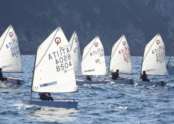 Flotta di piccole barche a vela Optimist con vele bianche e numeri velici, con giovani velisti a bordo, davanti a una costa rocciosa