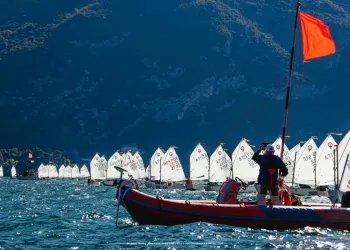 Flotta di Optimist allineati sul Lago di Garda con un gommone di supporto e bandiera rossa in primo piano