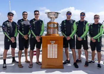 Sette uomini in divisa nero-verde posano accanto a un grande trofeo su una banchina con barche sullo sfondo.