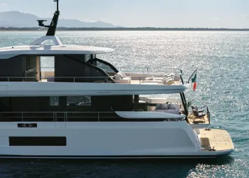 Yacht Azimut bianco in navigazione, vista laterale della poppa con terrazze, flybridge e bandiera italiana, su mare calmo con costa sullo sfondo