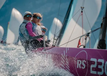 Equipaggio a bordo di un monotipo RS in regata con spruzzi d'acqua e vele sullo sfondo