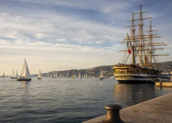 Nave a vela storica ormeggiata con numerose barche a vela in rada sullo sfondo.